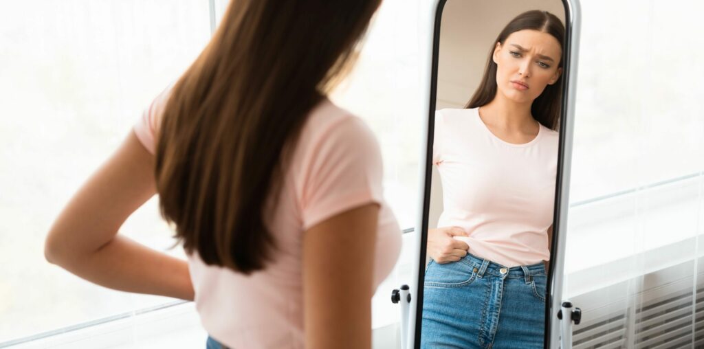 Traurige junge Frau misst Taille nach Gewichtszunahme, die zu Hause vor einem Spiegel steht