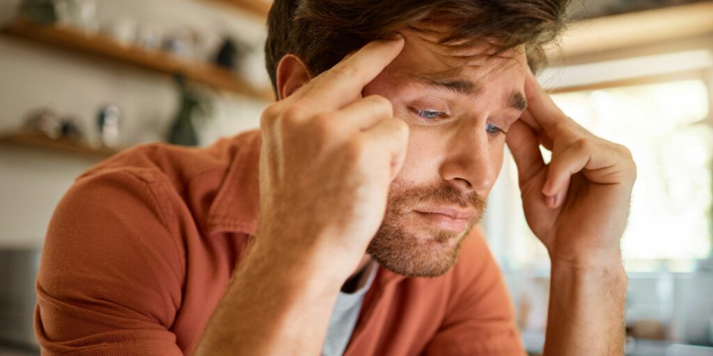 Junger Mann leidet unter Kopfschmerzen, während er allein von zu Hause aus arbeitet. Ein gestresster Mann sieht unglücklich aus, während er zu Hause arbeitet. Person sieht verärgert und besorgt aus